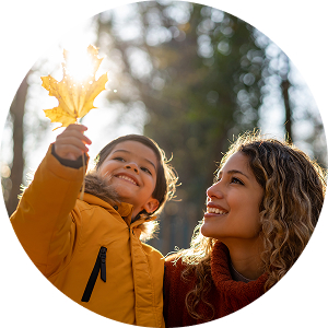 Smiling little boy holding a yellow leaf next to a smiling woman, with trees in the background.